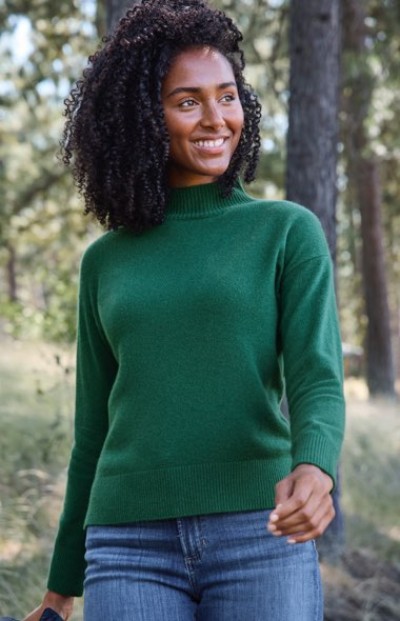 Person in green sweater and blue jeans walking outdoors in a forested area with trees in the background.