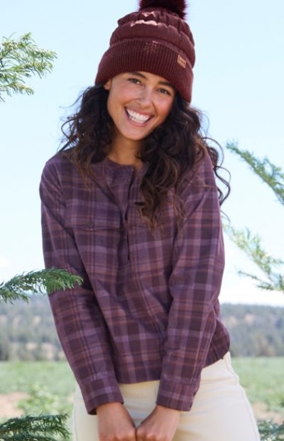 Person in maroon beanie and purple plaid shirt stands outdoors, smiling with trees and scenic background.