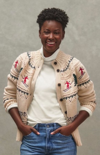 Person in beige cardigan with red skier design, white shirt, and blue jeans stands against a gray background.