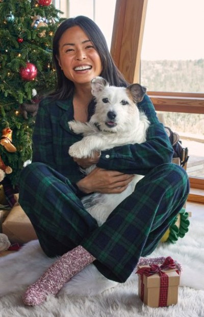 Person in plaid pajamas sits on white rug holding a dog, with a Christmas tree and wrapped gift nearby.