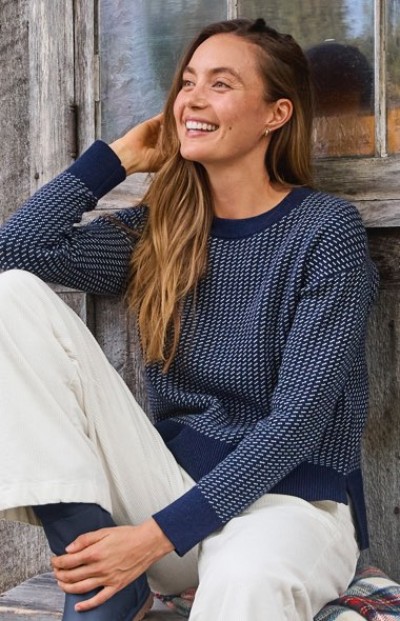 Person in blue patterned sweater and white pants sits by a rustic wooden wall with a window.