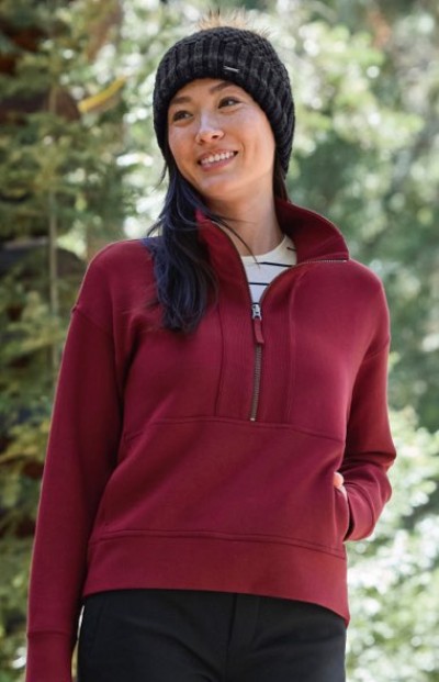 Person outdoors in maroon zip-up sweater and black knit hat, with greenery in the background.