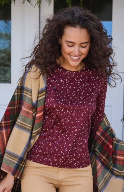Person with curly hair in maroon patterned shirt, beige pants, and plaid shawl stands in front of a door.