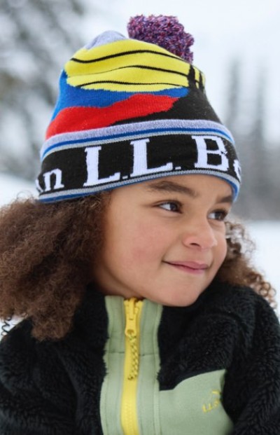Child in colorful hat and black jacket with yellow zipper, outdoors in a snowy setting.