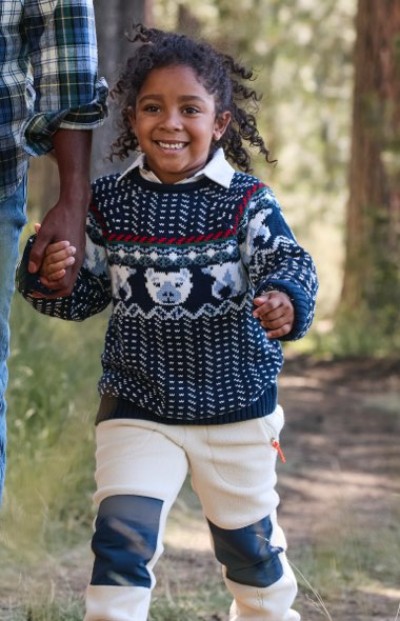 Child in patterned sweater and pants holds an adult's hand while walking outdoors.