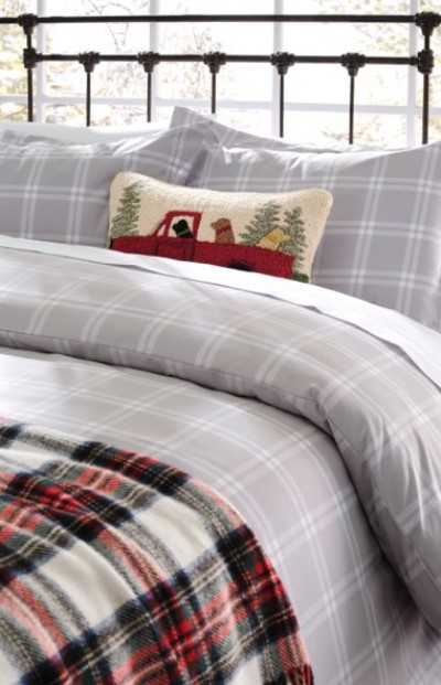 Bed with gray plaid bedding, red plaid blanket, and a pillow showing a red truck carrying trees for a festive touch.