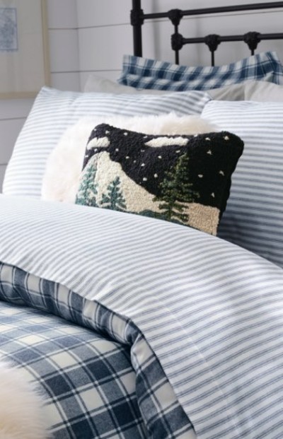 Cozy bed with blue striped and plaid bedding, topped with a pillow featuring a winter forest scene.