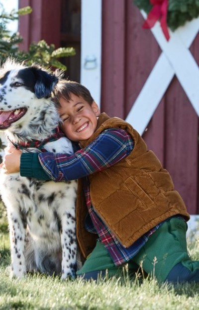 A child in a plaid shirt and brown vest hugs a black-and-white dog outside a red barn with white trim.