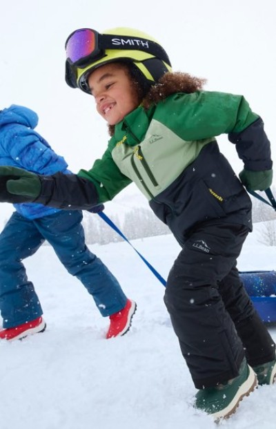 Two children in winter gear, one pulling a sled, enjoy a snowy outdoor activity.