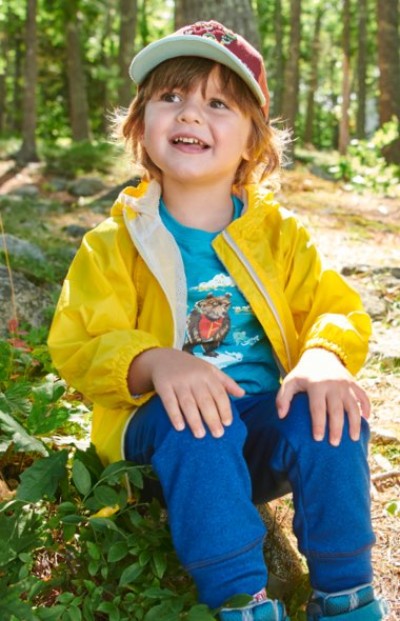 A child in a yellow jacket and blue pants sits outside on a sunny day, surrounded by greenery and trees.