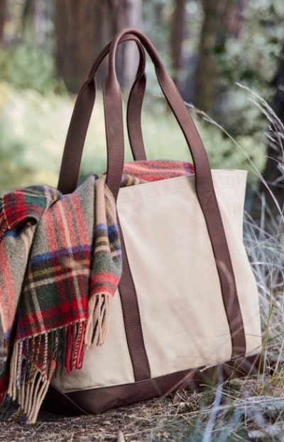 Beige tote with brown handles and a plaid blanket draped over it, placed outdoors in a forested setting.