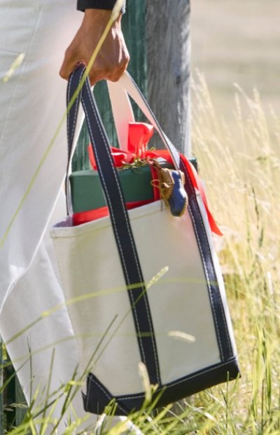 Person outdoors carrying a white tote bag with black straps containing a green gift box with red ribbon.