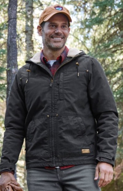 A person in a dark jacket, red plaid shirt, and orange cap stands in a forested outdoor setting.