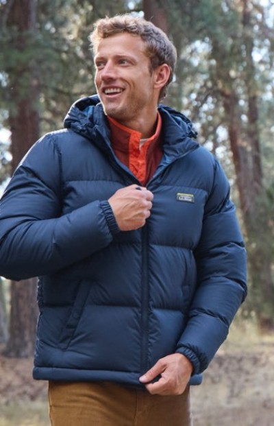 A person in a blue puffer jacket with a hood stands outdoors in a forested area.