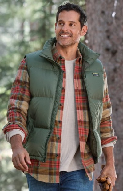 A person in a green puffer vest and plaid shirt holds gloves while standing outdoors with trees in the background.