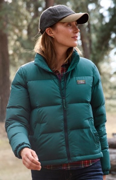 A person in a green puffer jacket and cap stands outdoors with trees in the background.