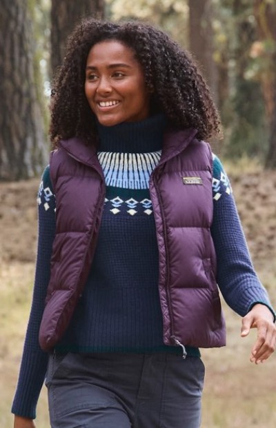 A person in a purple puffer vest and blue patterned sweater walks outdoors in a forested area.