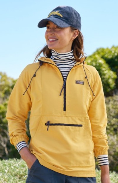 A person in a yellow jacket, striped turtleneck, and corduroy cap stands outdoors with greenery in the background.