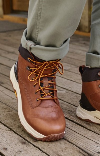 Brown hiking boots with orange laces on a wooden deck, worn with rolled-up light green pants.