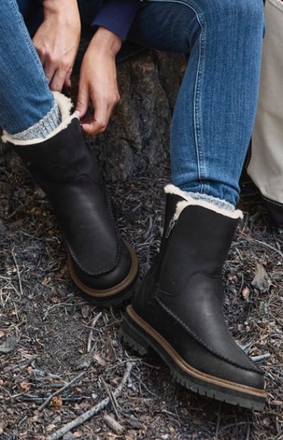 Person in black fur-lined winter boots sits on a log, adjusting the left boot in a cozy outdoor setting.