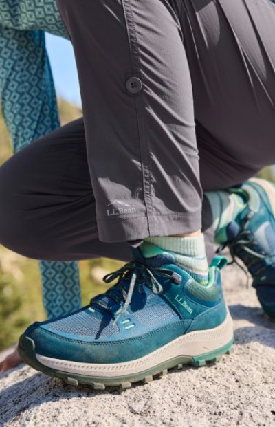 Person in gray L. L. Bean pants and teal hiking shoes kneels on a rock in an outdoor setting.