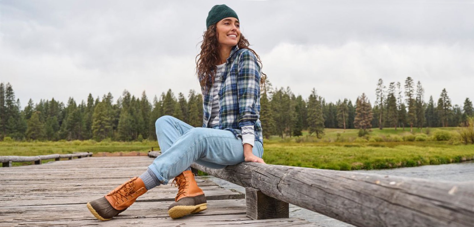 Woman sitting on a wooden dock in outdoor gear with boots, flannel shirt, and cap, near a grassy field and trees.