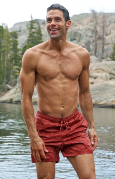 An athletic person standing in a lake wearing red swim shorts, with rocky terrain and pine trees in the background.