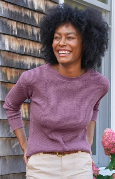Person in mauve textured crewneck sweater paired with light beige pants stands in front of a rustic shingled wall.