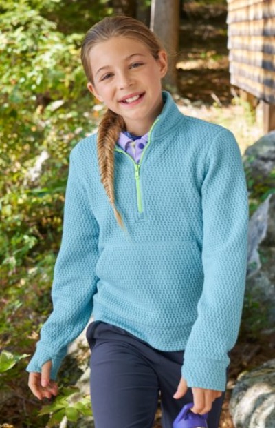 Child wearing a light blue textured pullover with a neon zipper and dark pants, standing outdoors near greenery and rocks.