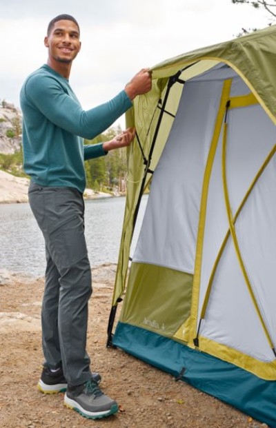 Person wearing a teal long-sleeve shirt and gray pants sets up a green and white tent near a lake in a forested area.