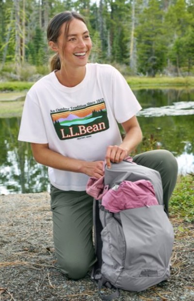 Person wearing an L.L.Bean graphic tee and green pants kneels by a pond, packing a gray backpack with a pink shirt.