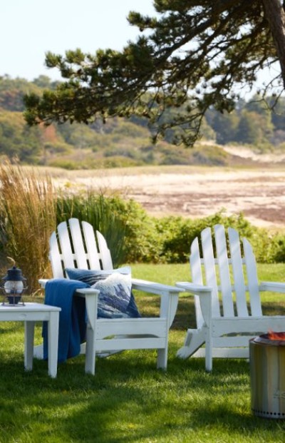 Two white Adirondack chairs with a blue throw and patterned pillow sit on a grassy lawn near a fire pit, overlooking a beach.