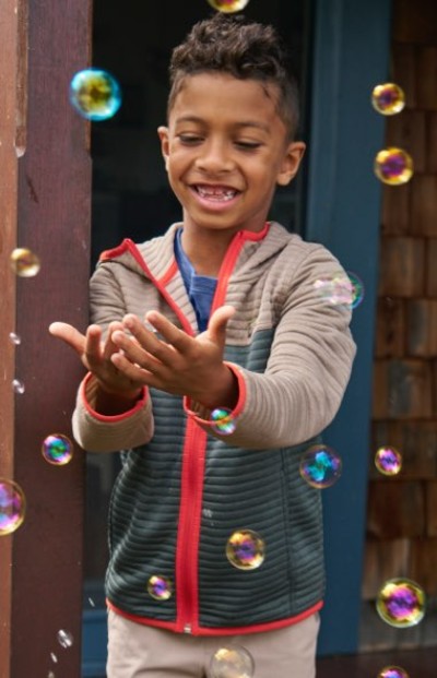 Child in a colorblock zip-up jacket with bright red trim, surrounded by floating bubbles in an outdoor setting.