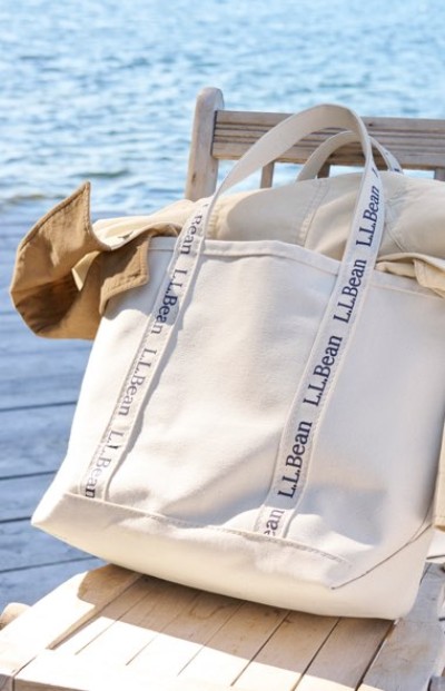 Large cream-colored L.L.Bean tote bag with branded handles sits on a wooden chair by the water, holding a tan jacket.