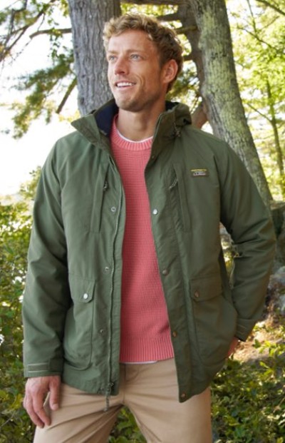 A person in a green outdoor jacket layered over a coral sweater, paired with beige pants in a forest setting.
