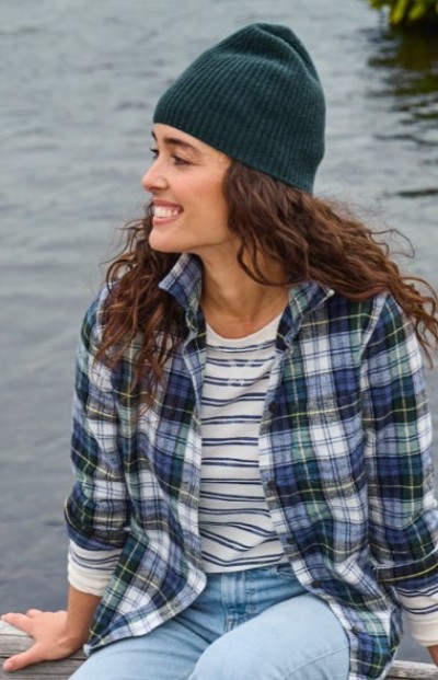 A smiling woman in a knit beanie and plaid flannel shirt sits outdoors by the water.