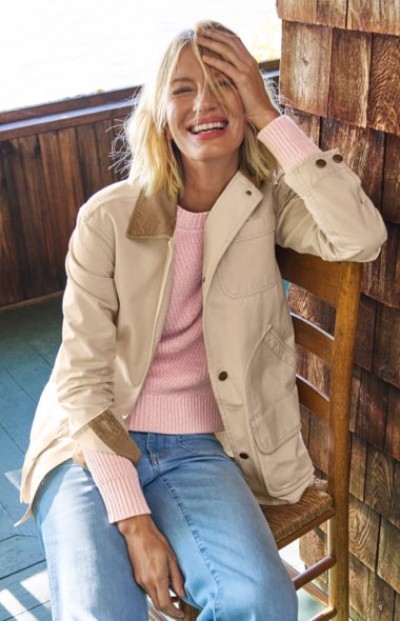 Woman in a beige jacket with brown accents layered over a pink sweater, in a rustic porch setting.