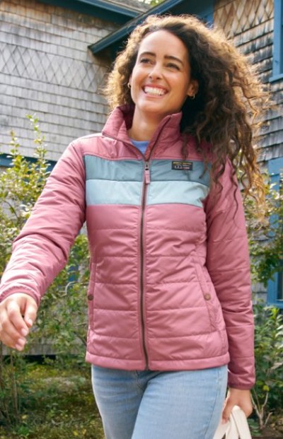 A person in a pink zip-up jacket with teal chest accents, styled for an outdoor casual look near a shingled building.