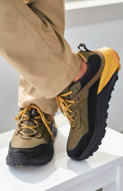 Olive and black hiking shoes with yellow accents and laces, paired with beige pants, resting on a white surface.