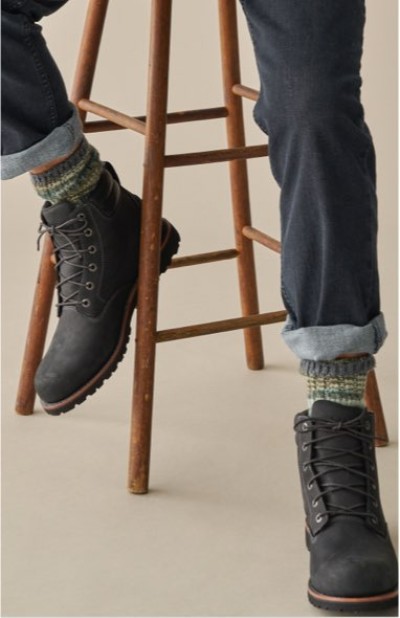 Black lace-up boots paired with rolled dark jeans and patterned socks, resting on a wooden stool.