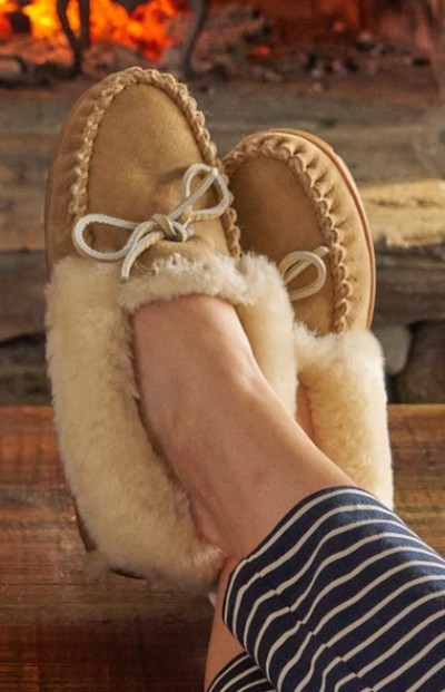 Tan shearling-lined moccasin slippers with white bows, worn with navy and white striped pants, resting near a cozy fireplace.