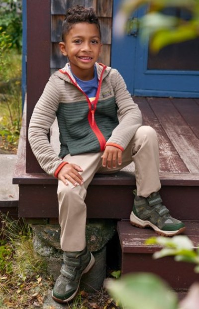 Child wearing a striped zip-up jacket, beige pants, and green hiking boots sits on wooden porch steps outdoors.
