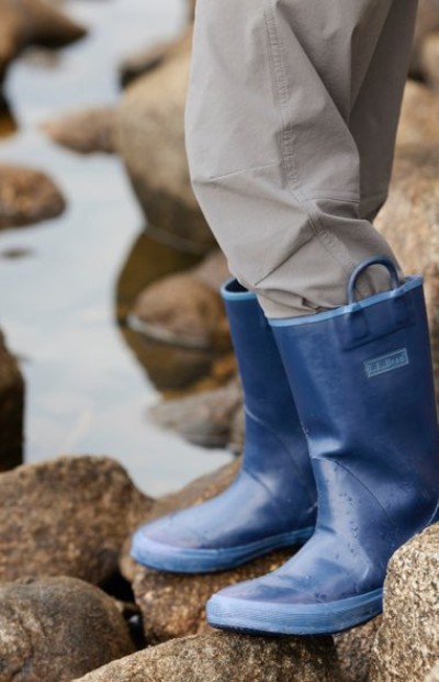 Blue waterproof rubber boots with loop handles are worn with light gray pants while standing on wet rocks near water.