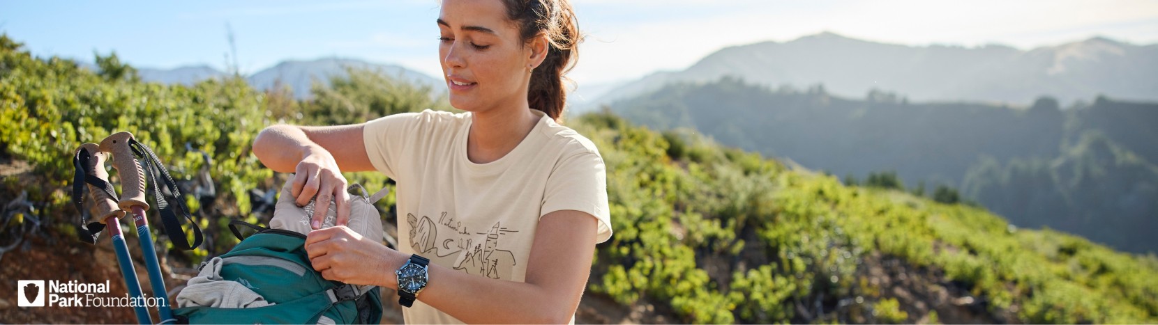 A woman wearing a beige shirt, outdoors on a grassy hill, opening a backpack. National Park Foundation logo in the corner