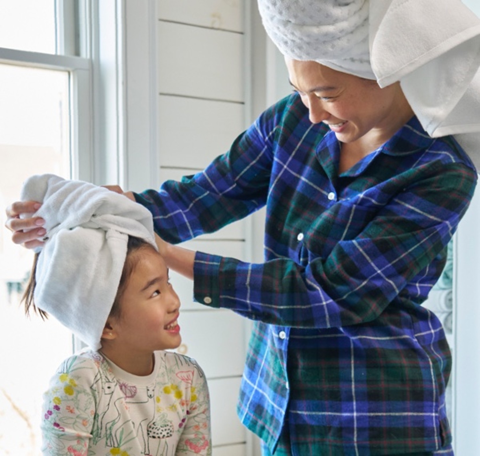 A woman wearing flannel pajamas with her hair wrapped in a towel, adjusts the towel wrapped around the head of a young girl.