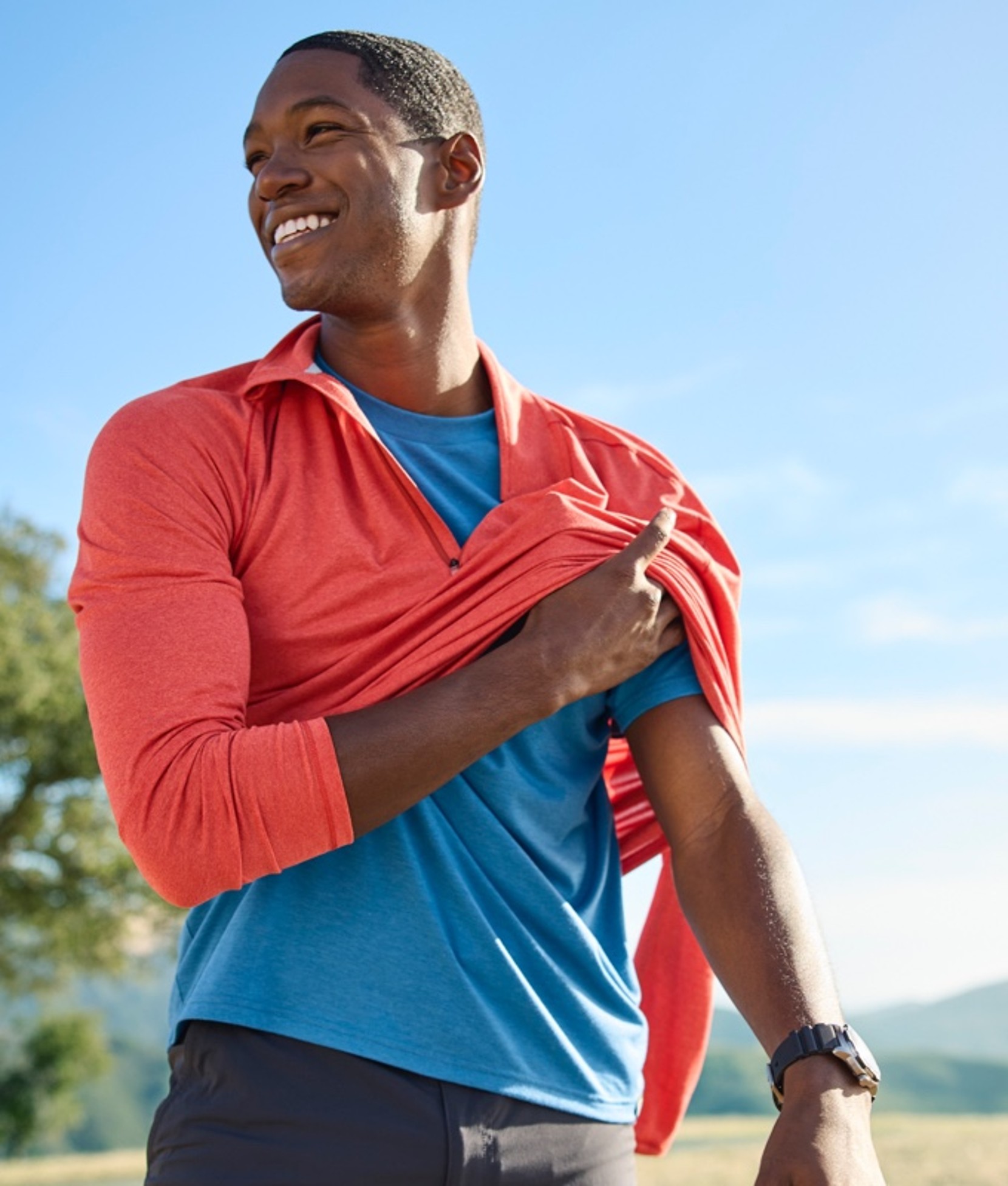 A smiling man outside, removing a long-sleeve layer.