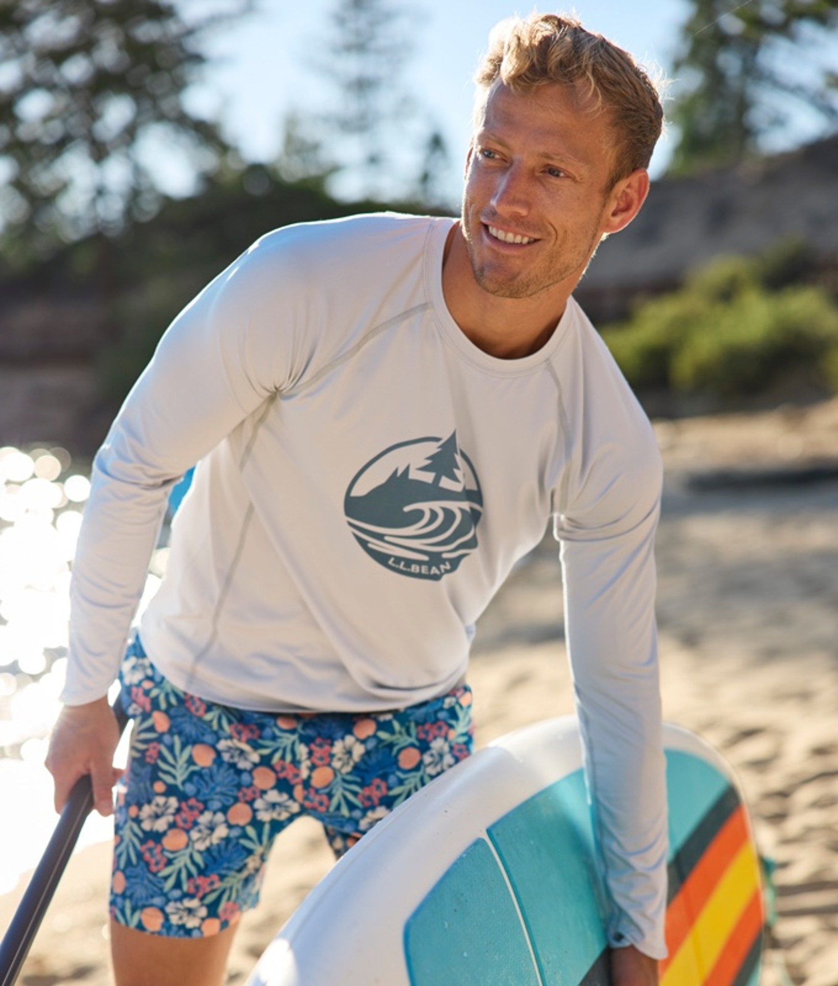 A man wearing swim trunks holding a stand-up paddleboard and paddle on the beach.