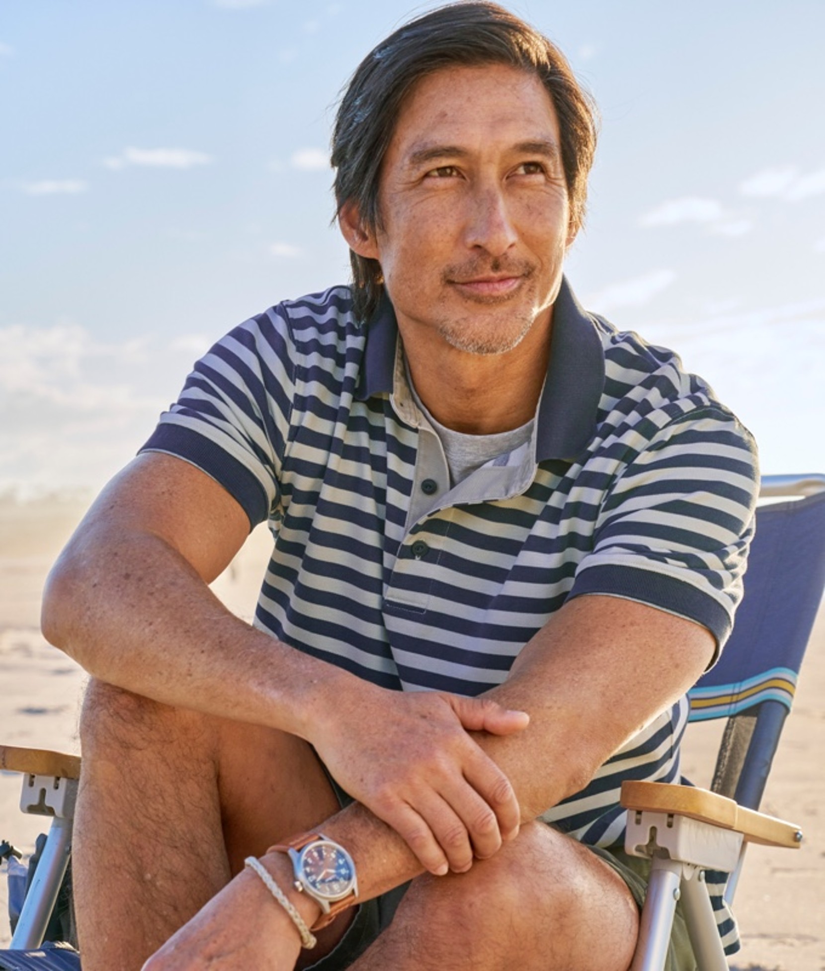 A man wearing a striped polo shirt sitting in a beach chair at the beach.