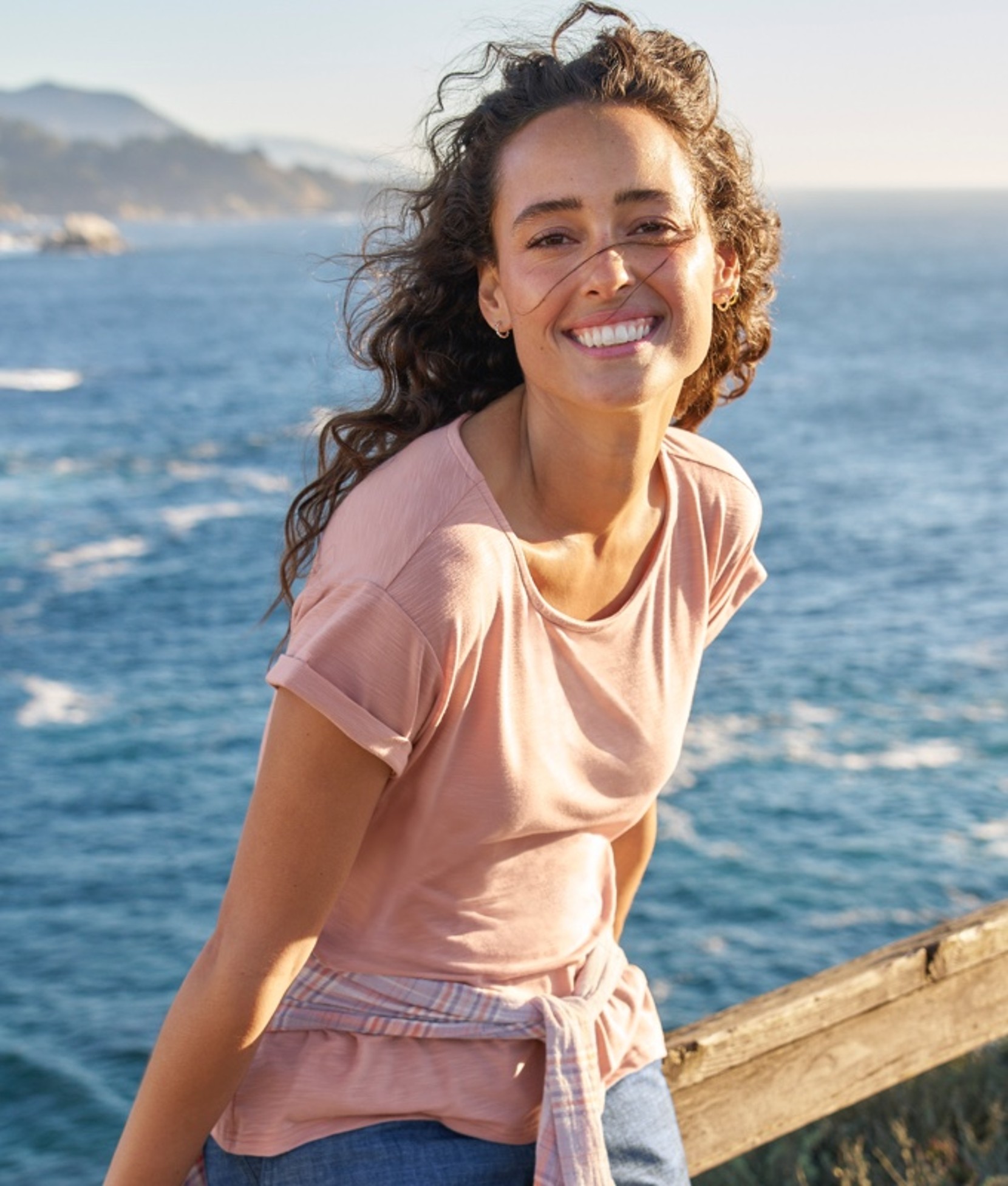 Smiling woman leaning on a dock railing, ocean waves in the background.