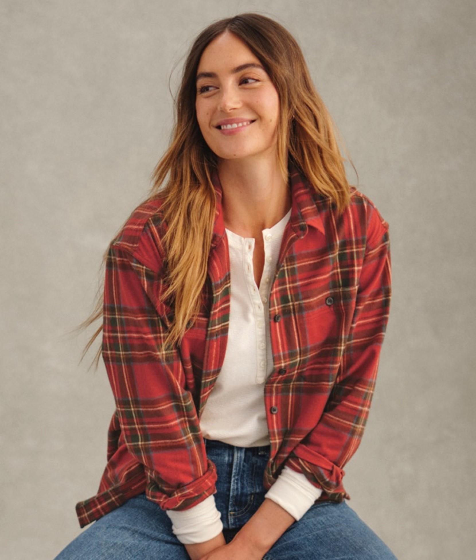 A women in red plaid flannel over white henley and blue jeans, seated.
